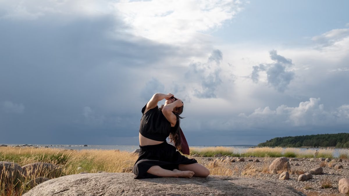 A woman performs a contemporary dance on a rock, surrounded by nature with a cloudy sky backdrop.
